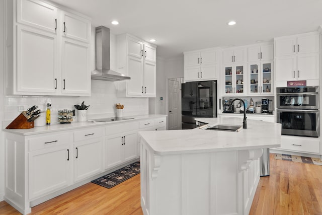 kitchen with an island with sink, sink, white cabinets, black appliances, and wall chimney range hood