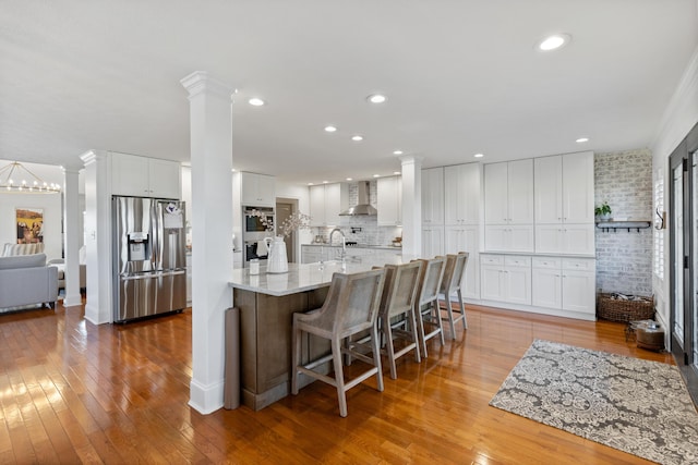 kitchen featuring wall chimney exhaust hood, light stone countertops, appliances with stainless steel finishes, white cabinets, and ornate columns
