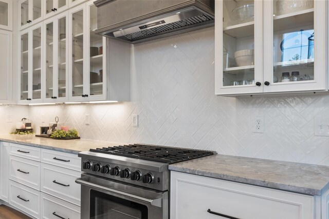 kitchen with light stone countertops, custom exhaust hood, stainless steel stove, white cabinetry, and backsplash