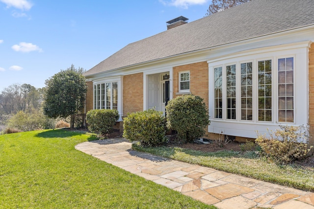 property entrance featuring a yard, brick siding, a chimney, and a shingled roof