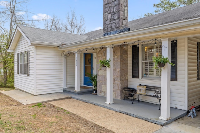 entrance to property featuring a porch, stone siding, roof with shingles, and a chimney