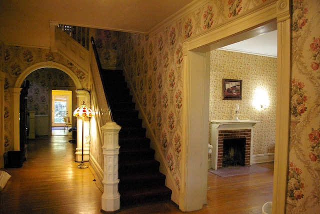 stairs with wallpapered walls, arched walkways, wood finished floors, and ornamental molding