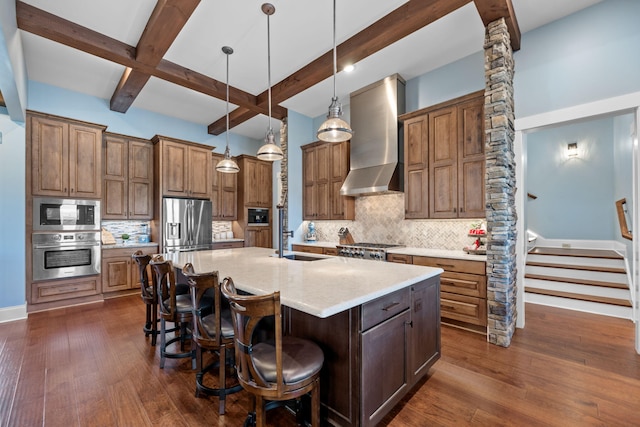 kitchen featuring wall chimney range hood, sink, appliances with stainless steel finishes, hanging light fixtures, and a center island with sink