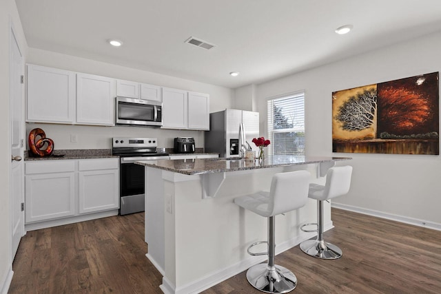 kitchen with dark wood-style floors, stainless steel appliances, visible vents, a kitchen island with sink, and dark stone countertops