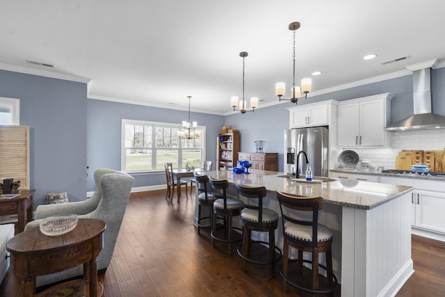 kitchen featuring appliances with stainless steel finishes, a kitchen island with sink, wall chimney range hood, decorative light fixtures, and a chandelier