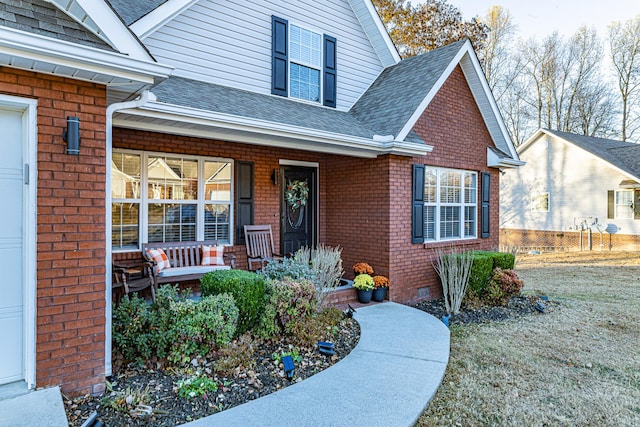 view of exterior entry with brick siding, covered porch, roof with shingles, and crawl space