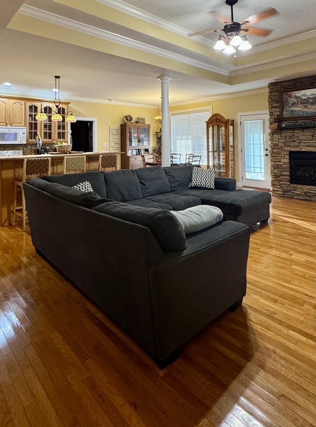 living room featuring a stone fireplace, hardwood / wood-style flooring, ornamental molding, ceiling fan, and a raised ceiling