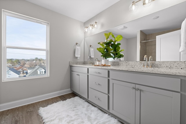 bathroom featuring double vanity, baseboards, wood finished floors, a tile shower, and a sink