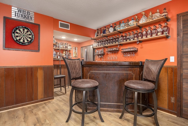 bar featuring stainless steel refrigerator with ice dispenser, wooden walls, light hardwood / wood-style flooring, and a textured ceiling