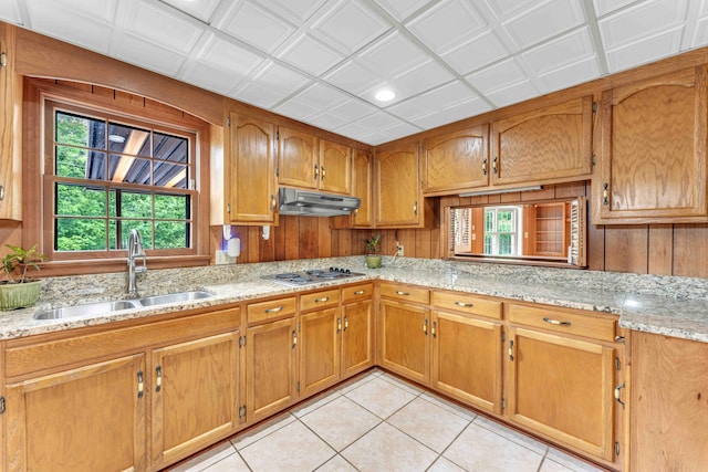 kitchen featuring light tile patterned floors, a sink, stainless steel cooktop, brown cabinetry, and under cabinet range hood