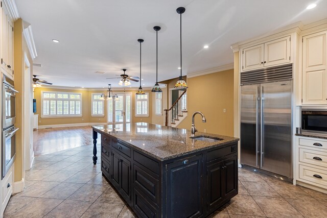 kitchen featuring hanging light fixtures, built in appliances, dark stone countertops, a center island with sink, and ornamental molding