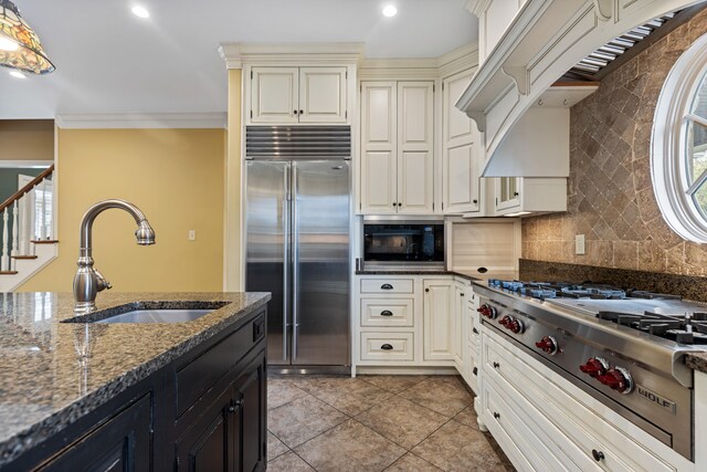 kitchen with sink, built in appliances, crown molding, dark stone counters, and light tile patterned flooring