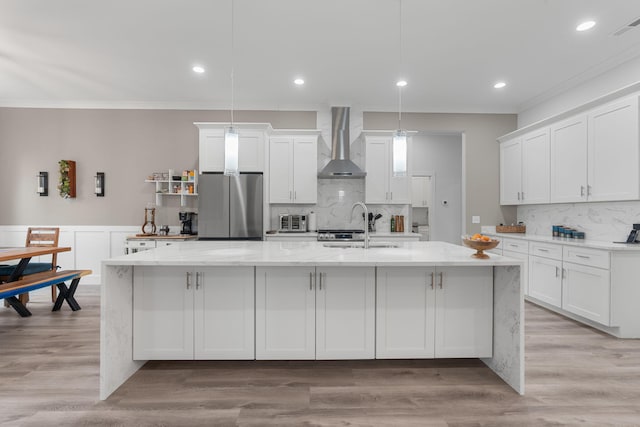 kitchen featuring wall chimney exhaust hood, hanging light fixtures, stainless steel fridge, a center island with sink, and white cabinets