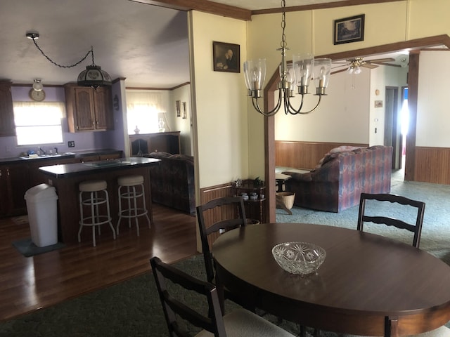 dining area featuring dark wood-style floors, wooden walls, wainscoting, and ceiling fan with notable chandelier