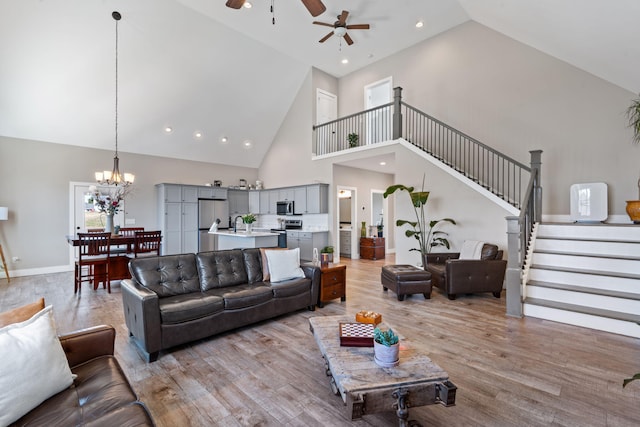 living room with baseboards, stairs, light wood-type flooring, high vaulted ceiling, and ceiling fan with notable chandelier