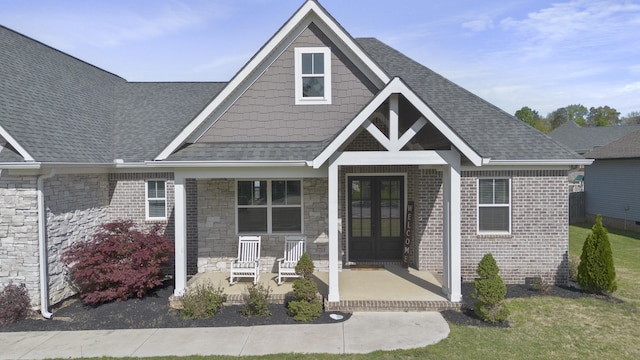 view of front of house with roof with shingles, a porch, stone siding, and french doors