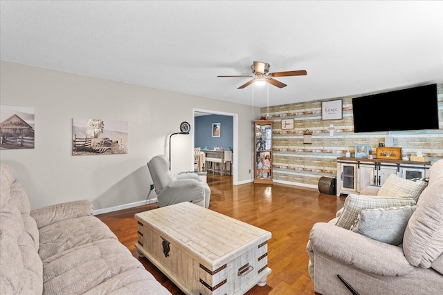 living room with ceiling fan, an accent wall, baseboards, and wood finished floors