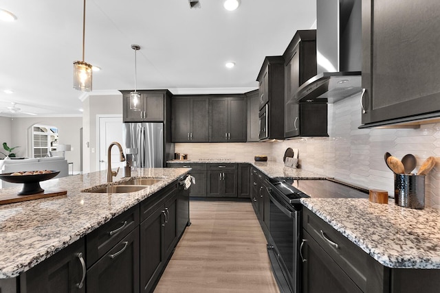 kitchen featuring wall chimney range hood, light wood-type flooring, light stone counters, appliances with stainless steel finishes, and a sink