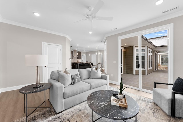 living area featuring crown molding, light wood-style flooring, ceiling fan with notable chandelier, and visible vents