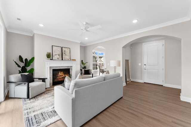 living room with light wood finished floors, crown molding, a fireplace, arched walkways, and a ceiling fan