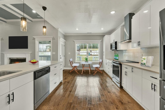 kitchen featuring wall chimney exhaust hood, hanging light fixtures, ornamental molding, stainless steel appliances, and white cabinets