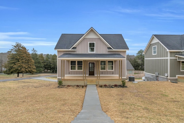 view of front facade with board and batten siding, roof with shingles, covered porch, a front lawn, and central AC