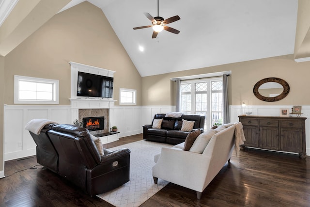 living area featuring dark wood-style floors, high vaulted ceiling, a high end fireplace, ceiling fan, and wainscoting