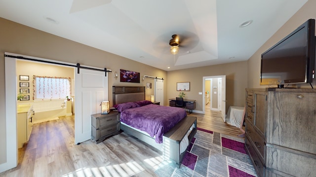 bedroom featuring ensuite bath, light hardwood / wood-style flooring, a raised ceiling, ceiling fan, and a barn door