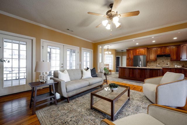 living room with french doors, light wood-type flooring, ornamental molding, ceiling fan with notable chandelier, and plenty of natural light