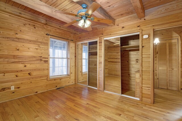 unfurnished bedroom with beam ceiling, multiple closets, ceiling fan, light hardwood / wood-style flooring, and wood walls