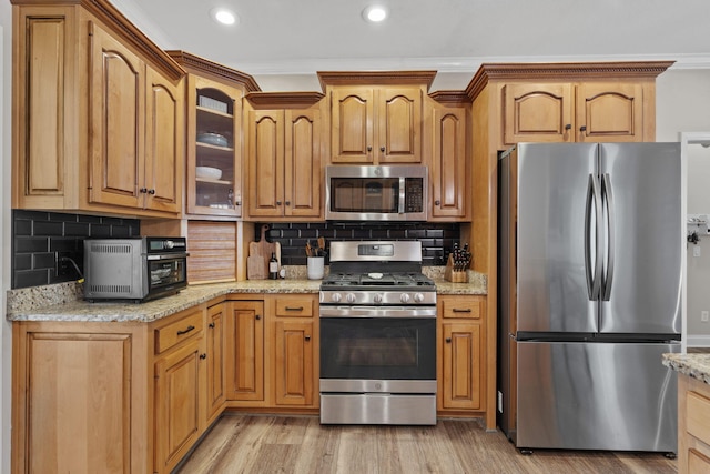 kitchen featuring stainless steel appliances, ornamental molding, light wood-type flooring, light stone countertops, and glass insert cabinets