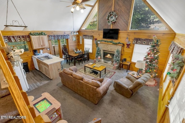 living room with ceiling fan, sink, high vaulted ceiling, a stone fireplace, and wood walls