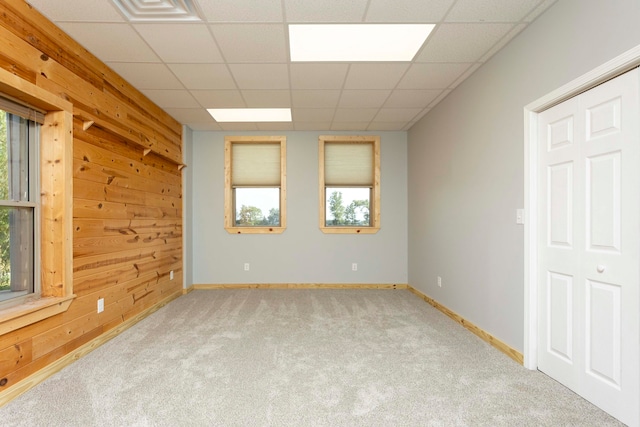 carpeted spare room with a drop ceiling, a healthy amount of sunlight, and wood walls