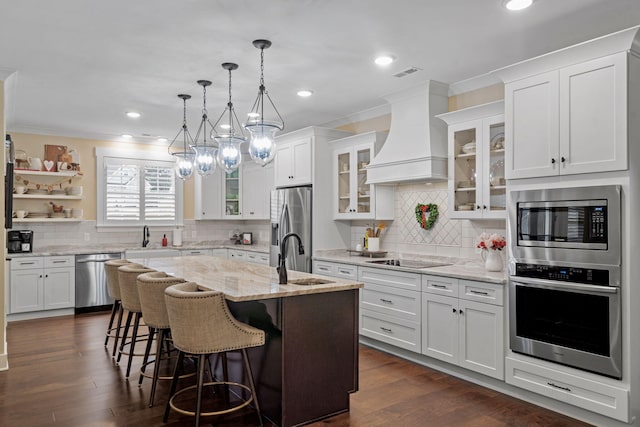 kitchen with pendant lighting, appliances with stainless steel finishes, a kitchen island with sink, custom range hood, and white cabinets