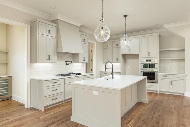 kitchen with beverage cooler, light wood-type flooring, a center island with sink, custom range hood, and appliances with stainless steel finishes