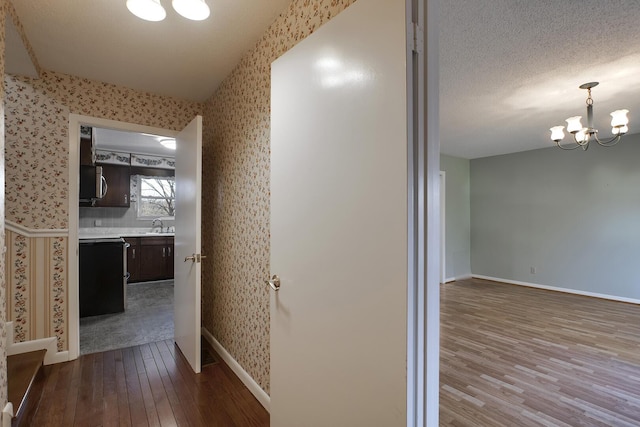 hall featuring a textured ceiling, wood finished floors, a chandelier, baseboards, and wallpapered walls