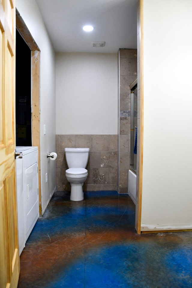 bathroom with shower / bath combination with glass door, tile walls, concrete floors, washer / clothes dryer, and toilet