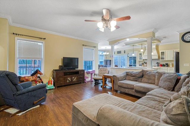 living room with dark wood-type flooring, crown molding, ceiling fan with notable chandelier, and decorative columns