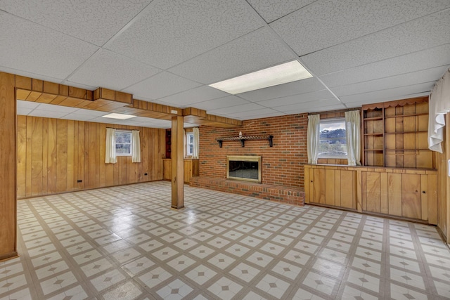 basement with built in shelves, wooden walls, a drop ceiling, and a brick fireplace