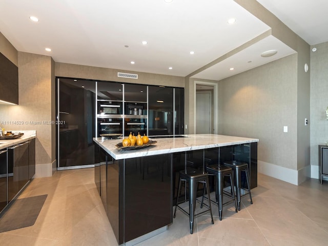 kitchen featuring double oven, a kitchen breakfast bar, light tile floors, a center island with sink, and light stone counters