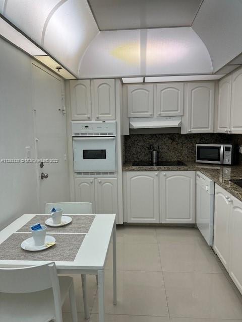 kitchen with backsplash, light tile flooring, white appliances, dark stone countertops, and white cabinets