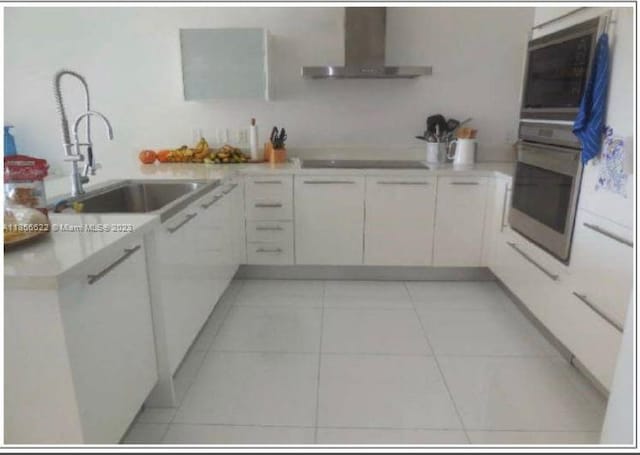 kitchen with light tile floors, oven, white cabinets, black electric cooktop, and wall chimney range hood