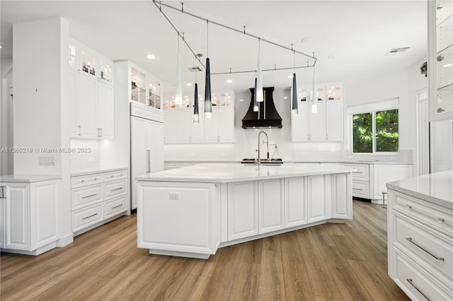 kitchen featuring an island with sink, white cabinets, light hardwood / wood-style floors, premium range hood, and light stone countertops