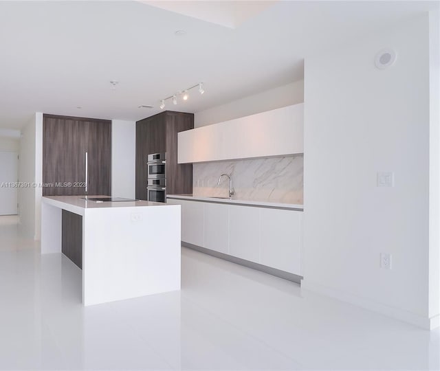 kitchen with stainless steel double oven, white cabinets, a center island with sink, and rail lighting