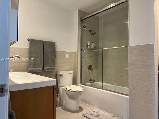 full bathroom with tile walls, toilet, vanity, and bath / shower combo with glass door