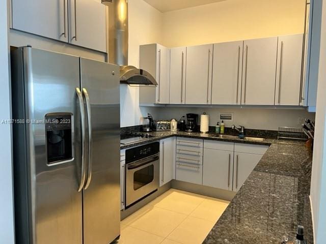 kitchen featuring sink, stainless steel appliances, light tile floors, dark stone countertops, and wall chimney exhaust hood