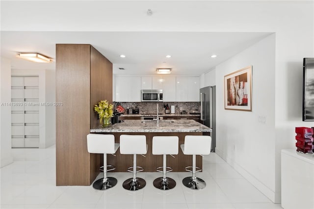 kitchen featuring light stone countertops, white cabinetry, backsplash, stainless steel appliances, and a breakfast bar area