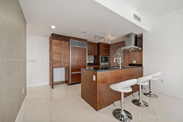 kitchen with built in appliances, wall chimney range hood, a kitchen bar, rail lighting, and tasteful backsplash