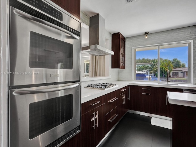 kitchen featuring dark tile patterned flooring, wall chimney range hood, sink, appliances with stainless steel finishes, and dark brown cabinetry