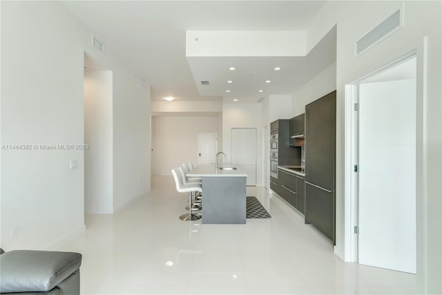 kitchen with light tile flooring, a kitchen bar, dark brown cabinetry, a kitchen island with sink, and sink